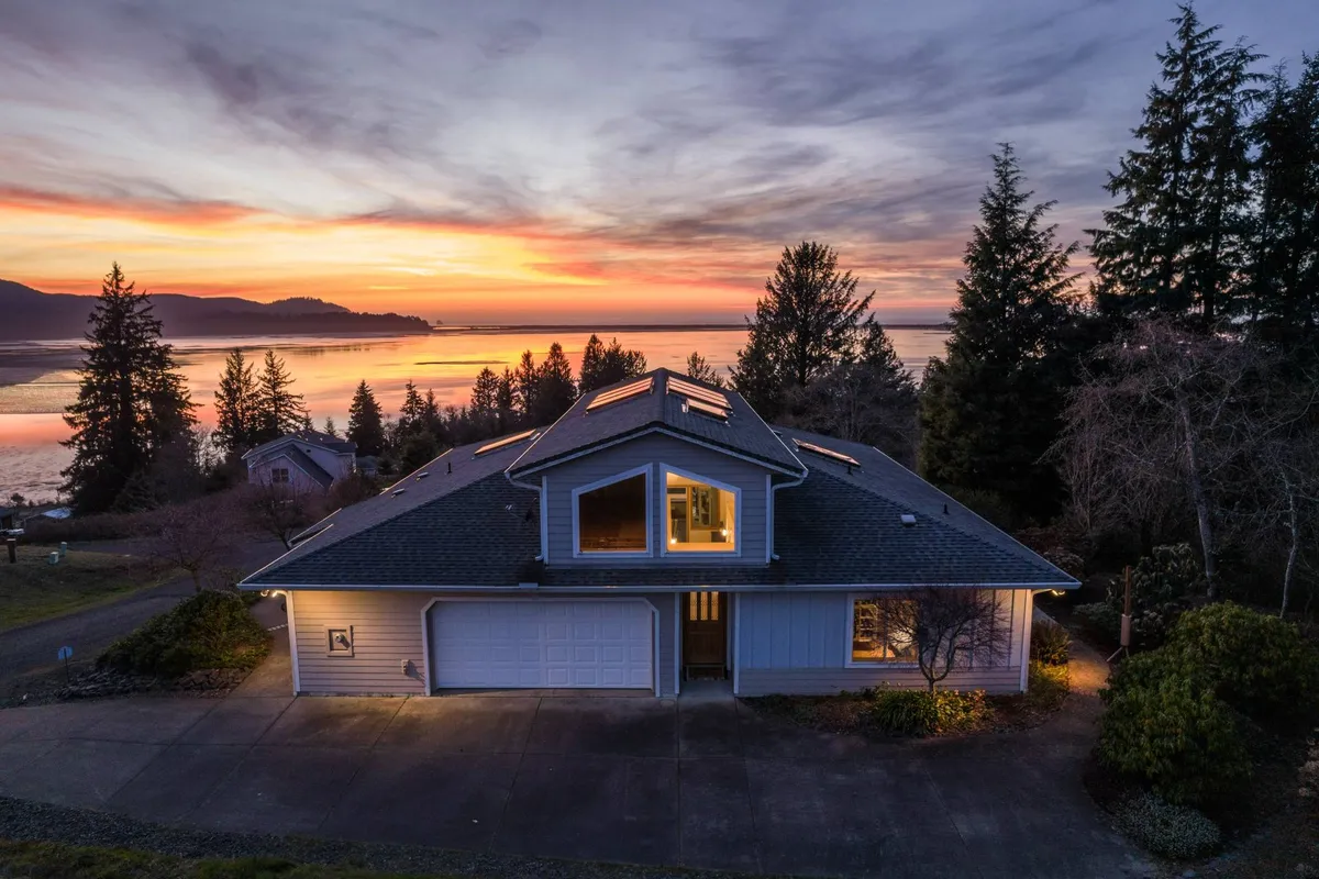 An exterior view of Bay View Villa at sunset, with soft sunlight reflecting off the water in front of the house. The villa is surrounded by lush trees, and the scene evokes a tranquil, inviting atmosphere. The expansive deck and porch are visible, hinting at the indoor-outdoor living experience.