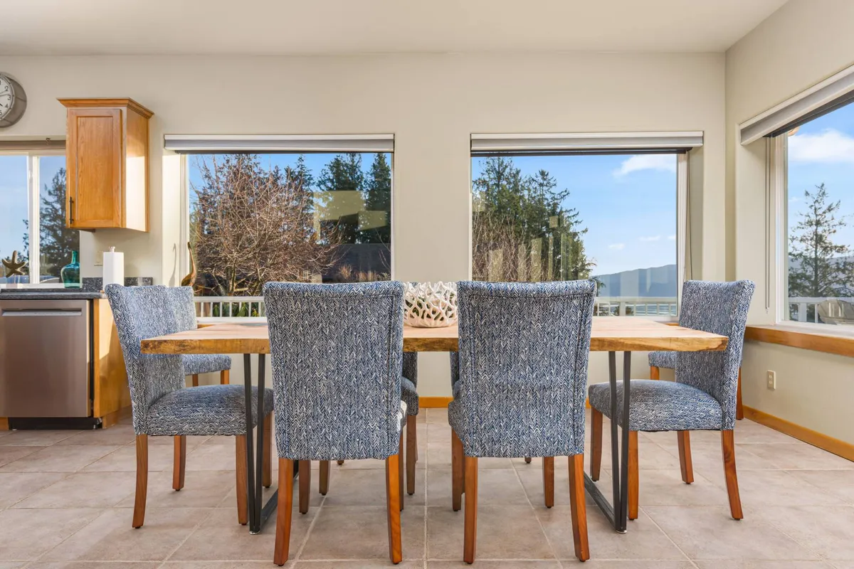 This image features a dining table placed near large windows that provide expansive views outside. The adjacent kitchen area is modern and inviting, with plenty of space for family meals and social gatherings. The windows suggest a strong connection between indoor living and the natural landscape beyond.