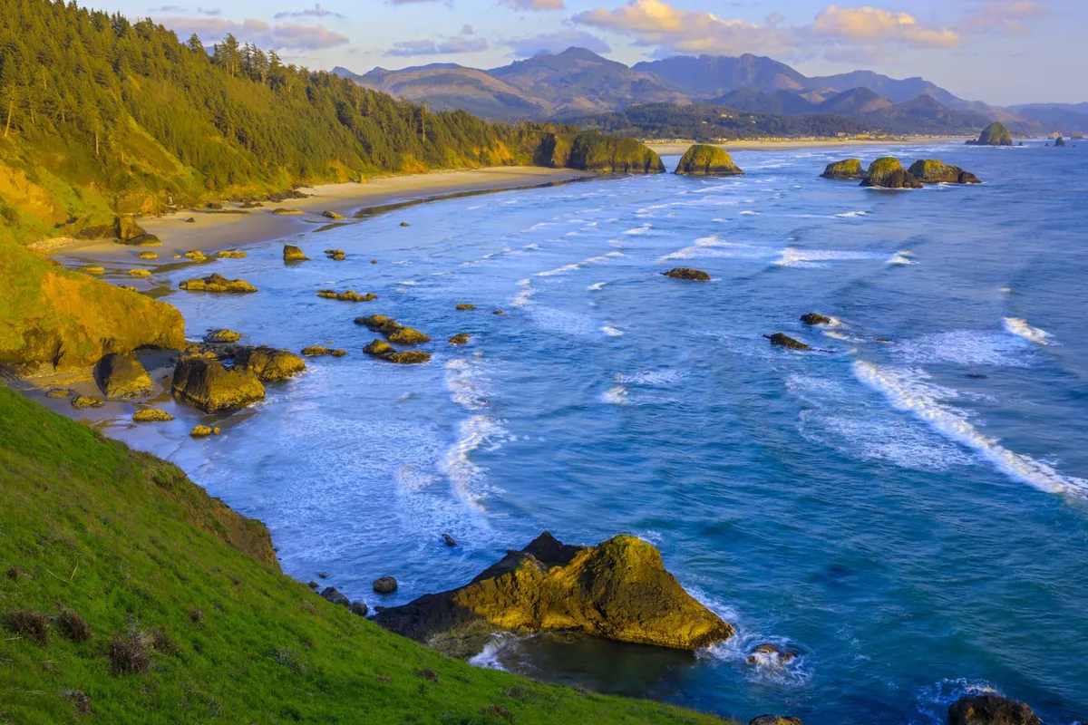 Featuring a vibrant view of the rocky Oregon coastline with waves crashing onto the sandy beach and distant mountains, this image captures the spirit of local beaches, such as Rockaway and Garibaldi, that are highlighted as pet-friendly destinations.