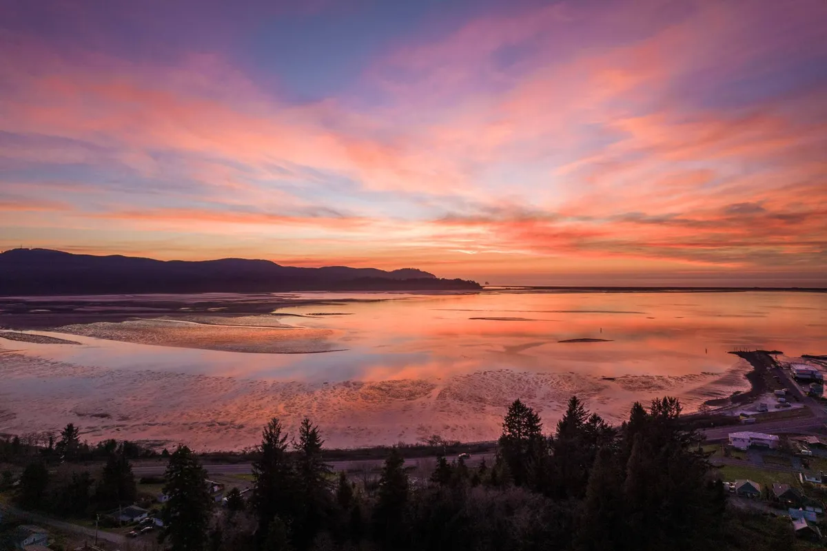 A stunning view of the sun setting over the water, with dramatic mountain silhouettes in the background and reflective ripples on the surface. The mood is serene and awe-inspiring, evoking moments of sunset watching and connection with Oregon's spectacular coast.