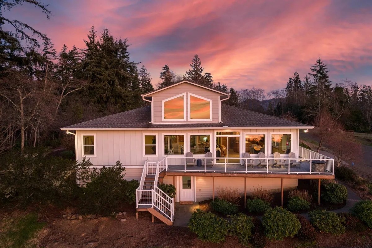 An exterior view of the Bay View Villa at sunset, featuring the house structure, balcony seating area, and the stunning sky ablaze with evening colors. The scene hints at outdoor gatherings and relaxation spaces overlooking beautiful views.