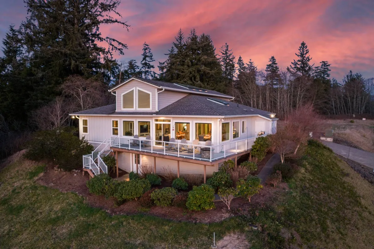 This exterior shot showcases the house’s spacious deck with large windows, surrounded by lush greenery and set against a sunset sky. The deck offers ample room for outdoor activities and gatherings, making it inviting for both people and pets.