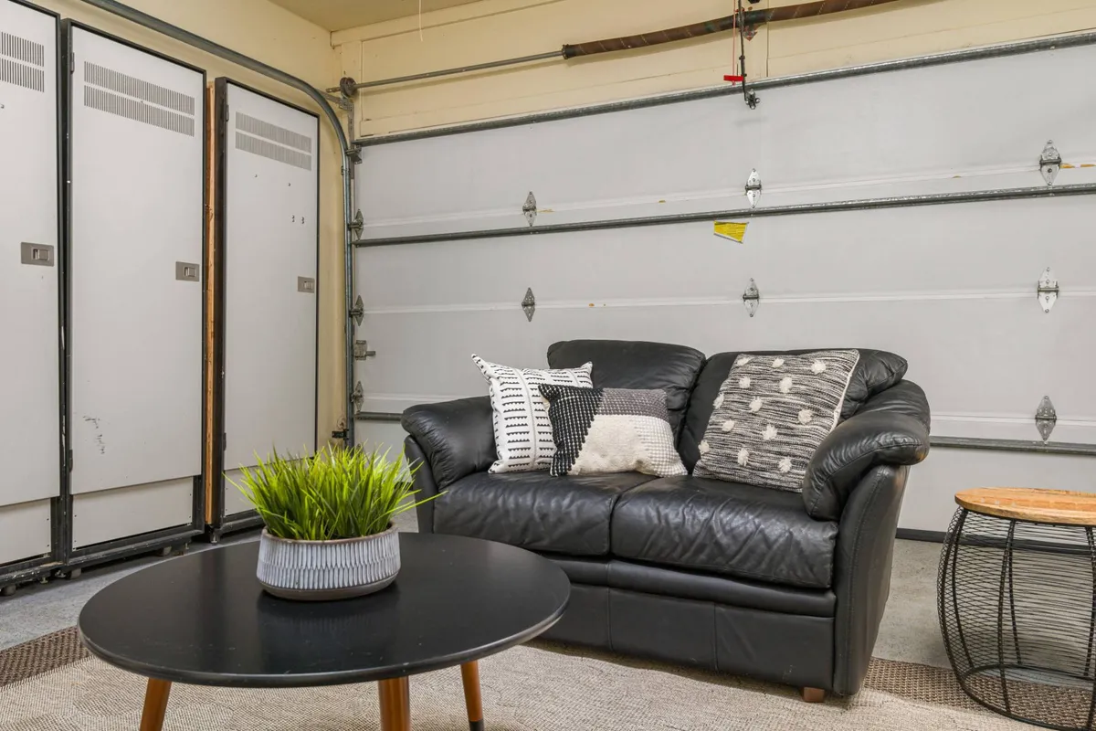 The image presents a comfortable interior seating area adjacent to a garage door, featuring a black leather sofa adorned with decorative pillows, a round coffee table, and a lush potted plant. The space looks bright and inviting, blending casual comfort with functional access, possibly near the hot tub area.