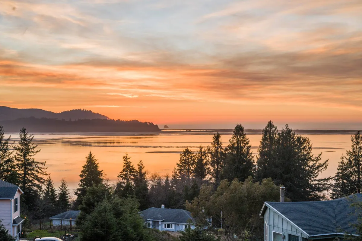 A wide, scenic vista featuring a vibrant sunset sky reflected on a calm water surface, with a distant mountain range and foreground trees. The mood is tranquil and awe-inspiring, evoking the magic of golden hour and the wild beauty of the Oregon coast.