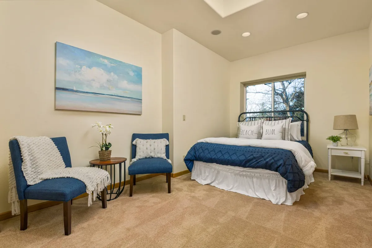 A serene bedroom featuring a bed with a blue quilt, inviting artwork on the wall, and a cozy seating area with chairs. The space is bathed in natural light, creating a restful atmosphere ideal for relaxation or quiet moments.