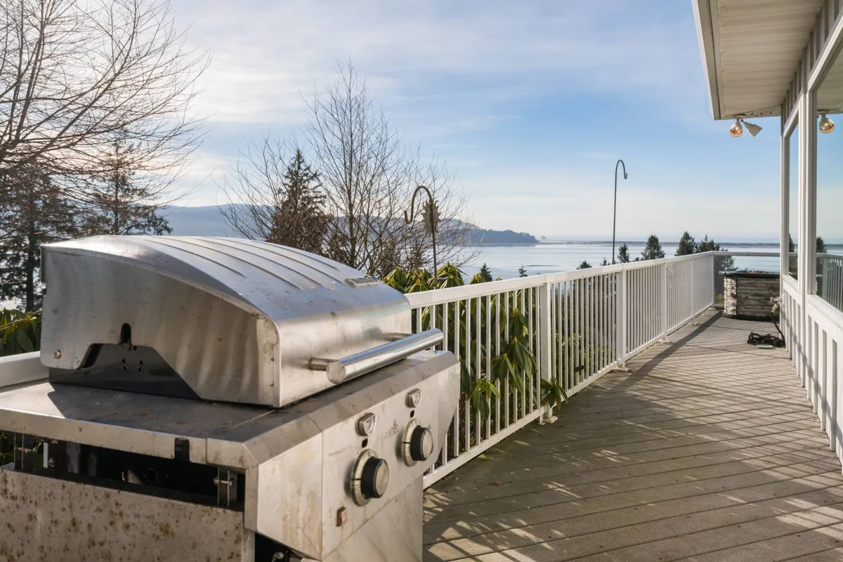 An inviting exterior deck scene featuring a grill, sturdy railing, and surrounding coastal trees, with a broad view overlooking the water. The scene is bathed in gentle natural light, suggesting either sunrise or sunset, and the setting evokes relaxation and appreciation of the outdoors.