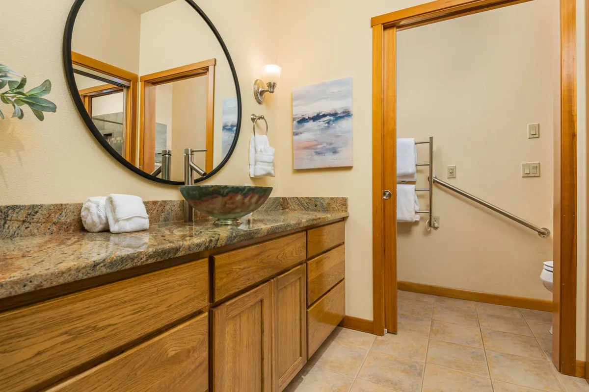 A serene bathroom with a granite countertop, circular mirror, tasteful artwork on the wall, and wooden cabinetry. The atmosphere is calm and spa-like, inviting relaxation and comfort.