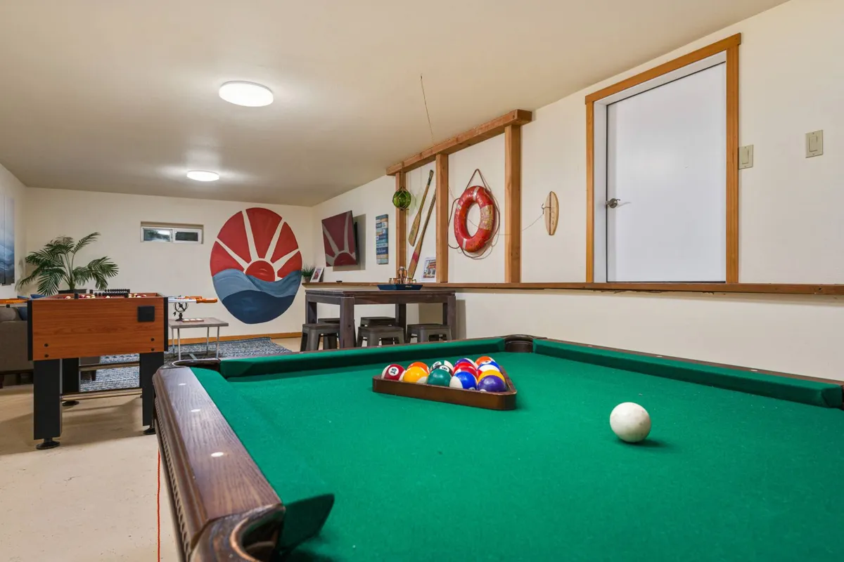 A lively game room featuring both a pool table and a foosball table, with a striking sunset mural on the wall and a dining table nearby. The space is bright and inviting, clearly set up for group activities and socializing.