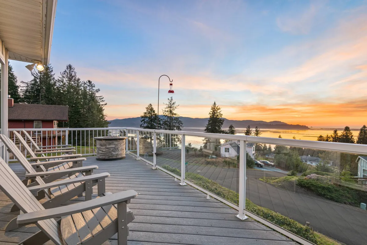 The image shows a spacious deck with comfortable chairs overlooking a breathtaking panorama of Tillamook Bay and the Pacific Ocean. The sky is painted with vibrant sunset hues, and lush trees frame the tranquil water view, creating a peaceful, inviting atmosphere.