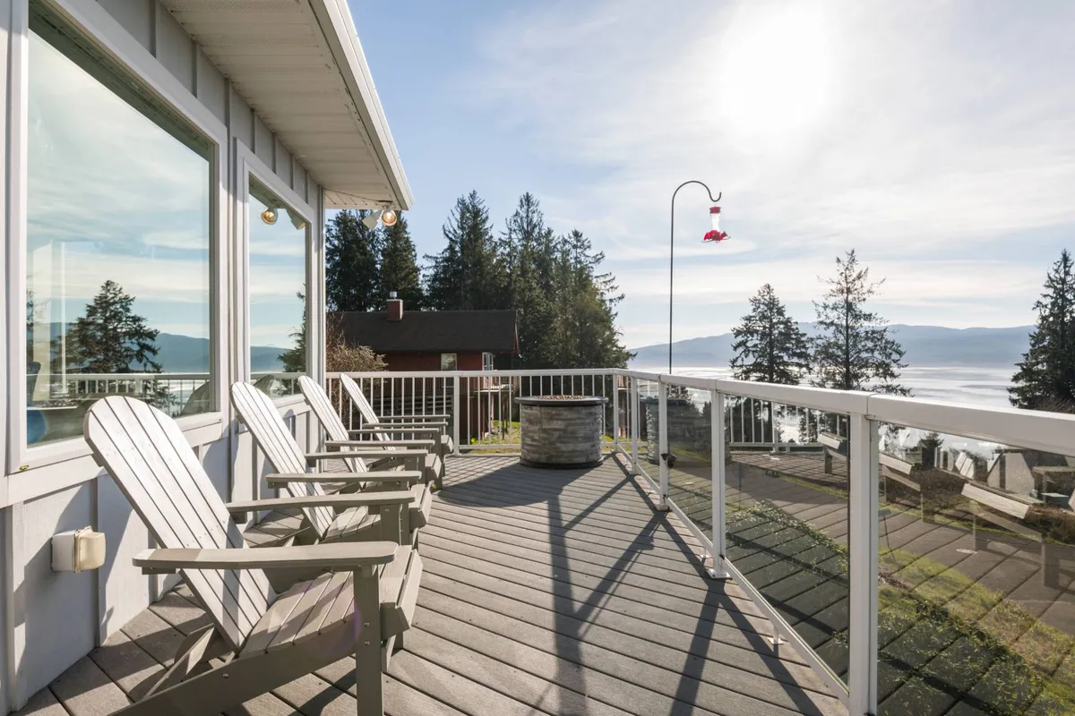 A peaceful lakeside or bayfront patio with Adirondack chairs positioned to face the water, a hummingbird feeder, and lush trees framing the view. The tranquil setting is ideal for birdwatching, quiet contemplation, or taking in sunsets and stargazing.