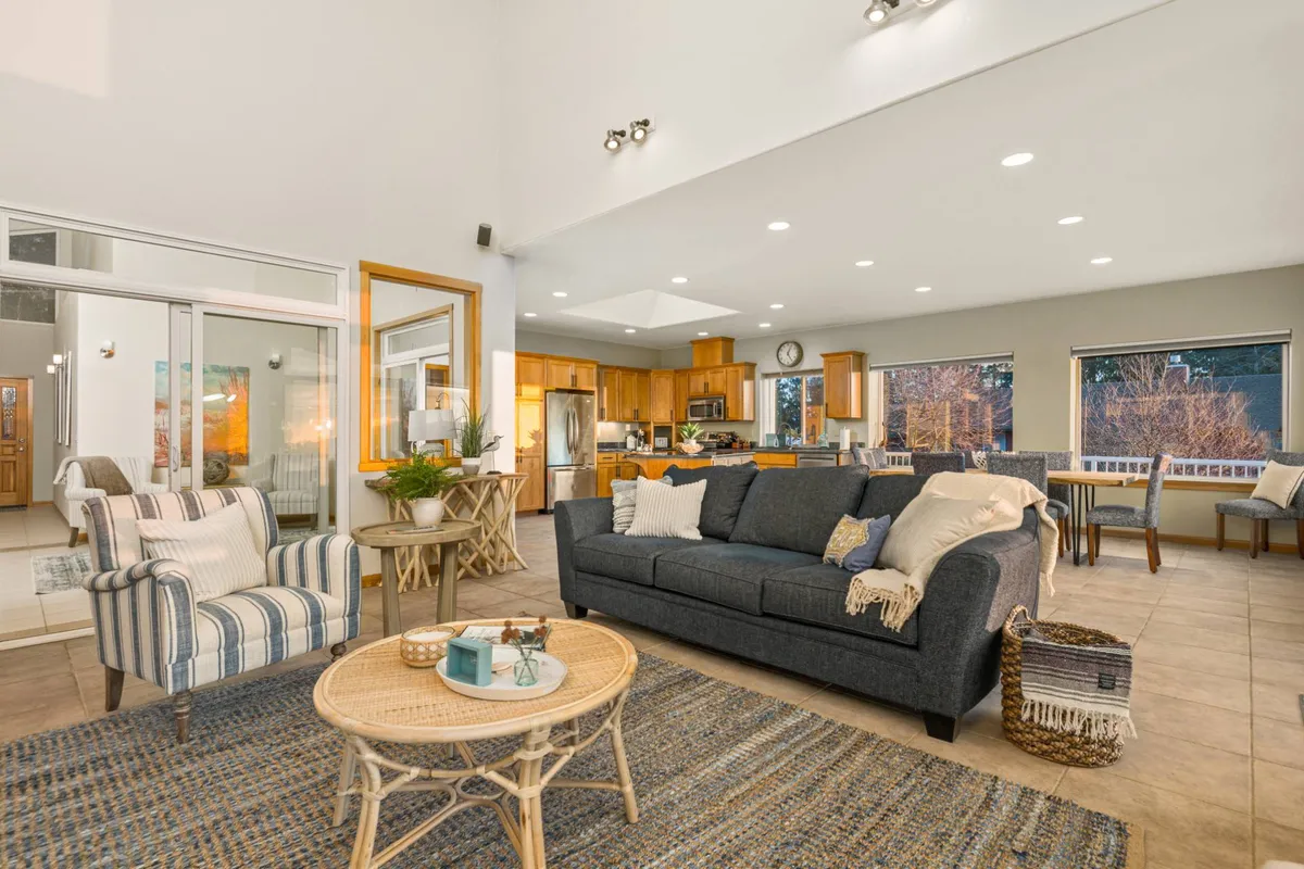 The image showcases an open-concept living space featuring a comfortable sofa, a coffee table, and a view of the kitchen and dining areas in the background. Large windows allow sunlight to flood the space and highlight the inviting ambiance. The design encourages social gatherings and relaxation.